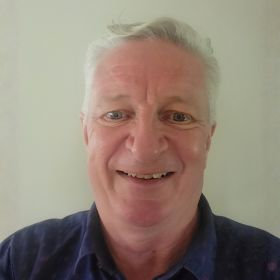 Smiling older man with gray hair wearing a dark shirt, looking at the camera against a light neutral background.