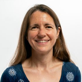 Dr Clare Ellis-Smith - DEMENTIA RESEARCHER Portrait of a smiling woman with long brown hair in a blue patterned sweater against a light background.