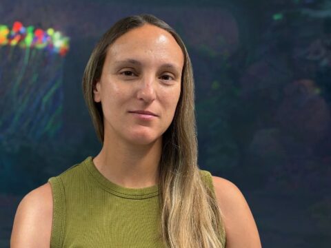 Portrait of a woman with long blond hair, wearing an olive sleeveless top, looking at the camera with a calm expression, aquarium background with colorful fish on the left