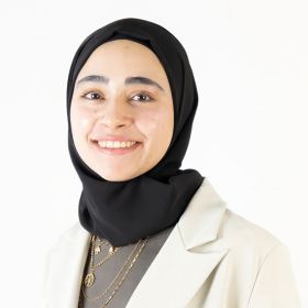 Abrar AbuHamdia - DEMENTIA RESEARCHER Smiling woman in a black hijab, beige blazer, and layered necklaces against a white background.