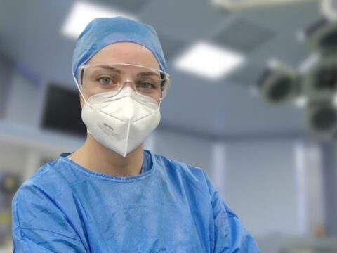 Medical professional in blue surgical scrubs, cap, mask, and protective goggles in an operating room