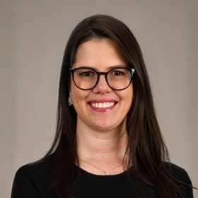 Portrait of a smiling woman with shoulder-length dark hair, black-framed glasses, wearing a black top.