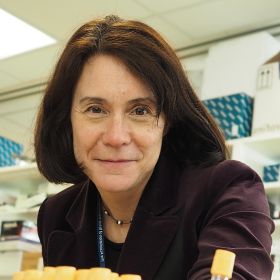 Professor Michele Hu - DEMENTIA RESEARCHER Woman in a burgundy blazer smiling at the camera in an office or lab workspace behind her shelves and supplies.