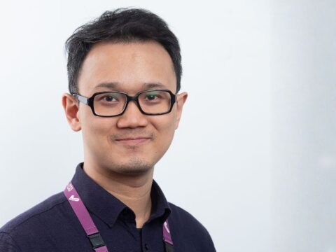 Portrait of a man with black glasses and a dark polo, smiling, wearing a purple lanyard.