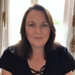 Emma Williams - DEMENTIA RESEARCHER Smiling woman with shoulder-length brown hair wearing a black top, indoors with daylight behind her.