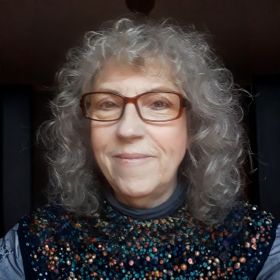 Sue Williams - DEMENTIA RESEARCHER Portrait of a smiling woman with gray curly hair and red-framed glasses, wearing a dark floral-patterned top.
