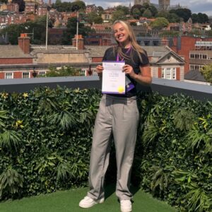 Claire Rowley Collecting an Award Claire Rowley standing outdoors holding her Una Holden Award 2025 certificate, with a cathedral and city buildings behind her.