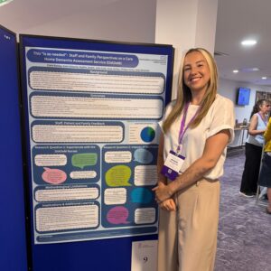 Claire Rowley Conference Poster Claire Rowley standing beside a research poster about staff and family perspectives of a care home dementia assessment service.