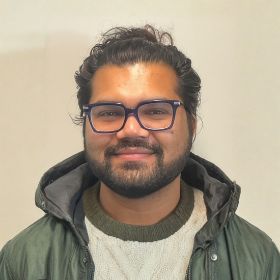 Dr Pulkit Khandelwal Portrait Dr Pulkit Khandelwal who is a bearded man with dark hair and beard, blue glasses, wearing a green jacket and sweater, smiling at the camera.