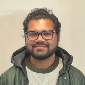 Dr Pulkit Khandelwal Portrait Dr Pulkit Khandelwal who is a bearded man with dark hair and beard, blue glasses, wearing a green jacket and sweater, smiling at the camera.