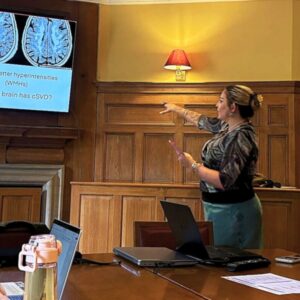 Honey Blossom McLaughlin pointing to a presentation screen showing brain scans in a wood-panelled room.