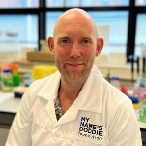 Patrick Lewis Close-up of a smiling man in a white lab coat with a name badge, in a bright indoor setting with blurred background.