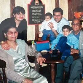 A family group seated and standing around a small restaurant table, with a wall plaque reading "IN HONOUR OF..." visible behind them. On the left, an older woman in a grey outfit sits with sunglasses pushed up into her hair, her hand resting on the table. Behind her, a younger woman with dark hair and sunglasses stands. To the right, a man in a striped top holds a baby, and in front of him a young boy in a blue outfit sits at the table. On the far right, an older man in a pale blue shirt sits smiling.