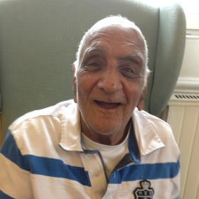 An older man with white hair sits smiling in a green armchair, wearing a white polo shirt with blue stripes. The setting, with its plain furnishings and armchair, suggests a care home environment.
