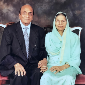 A formal studio portrait of an older man and woman seated side by side against a plain grey backdrop. The man wears a dark suit jacket, striped shirt and tie, hands resting on his lap. The woman wears a pale mint green sari with a matching dupatta covering her head, hands folded in her lap.