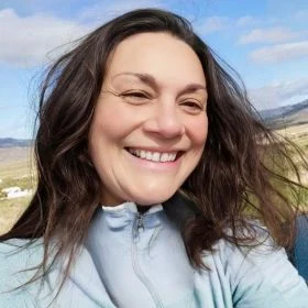 Woman smiling at the camera outdoors in a light blue zip-up jacket with mountains and a blue sky behind her.