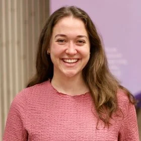 Smiling woman with long brown hair wearing a pink textured sweater, posed against a purple wall."
